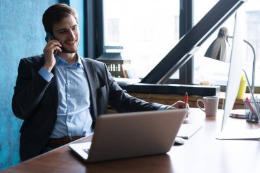 Ofis masasında dizüstü bilgisayarıyla oturan ve cep telefonuyla konuşan yakışıklı genç bir erkek portresi. İletişim konsepti.