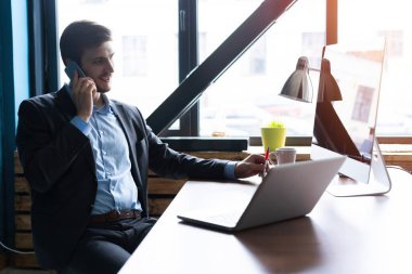 Ofis masasında dizüstü bilgisayarıyla oturan ve cep telefonuyla konuşan yakışıklı genç bir erkek portresi. İletişim konsepti.