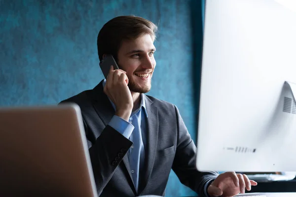 Portret van een knappe jonge man die achter een bureau zit met een laptop en praat op een mobiele telefoon. Communicatieconcept. — Stockfoto
