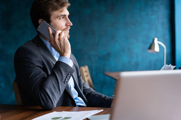 Porträt eines hübschen jungen Mannes, der mit Laptop am Schreibtisch sitzt und mit dem Handy telefoniert. Kommunikationskonzept. — Stockfoto