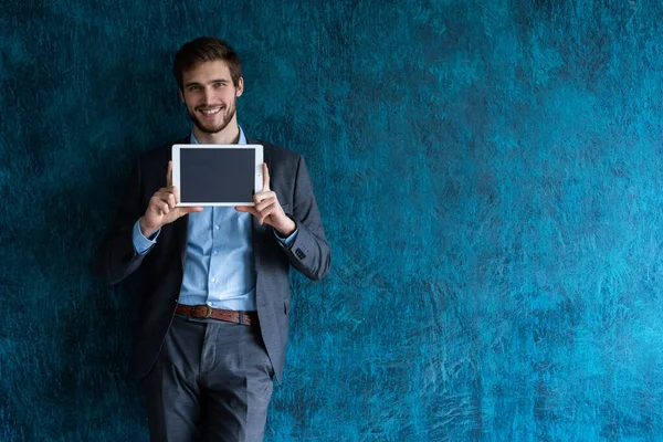 Uśmiechnięty młody biznesmen na niebieskim tle w klasycznym szarym garniturze prezentujący tablet. — Zdjęcie stockowe