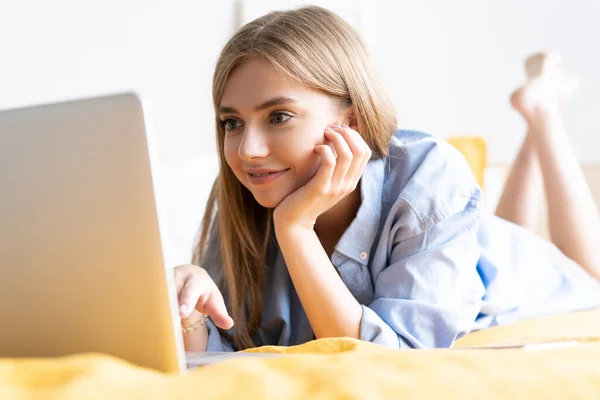 A smiling woman lying down the bed in front of her laptop. Working from home in quarantine lockdown — Stock Photo, Image