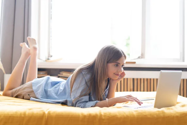 A smiling woman lying down the bed in front of her laptop. Working from home in quarantine lockdown — Stock Photo, Image