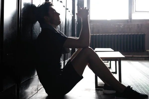 Ung, atletisk, hvit mann som sitter alene i en mørk garderobe og hviler etter trening. – stockfoto