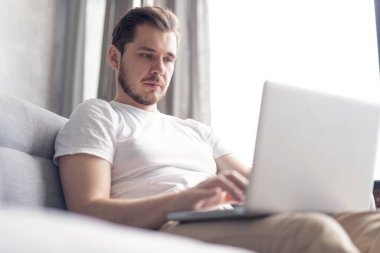 Yeni blog yazıları yazılıyor. Yakışıklı genç adam evdeki kanepede otururken bilgisayarını gülümseyerek kullanıyor..