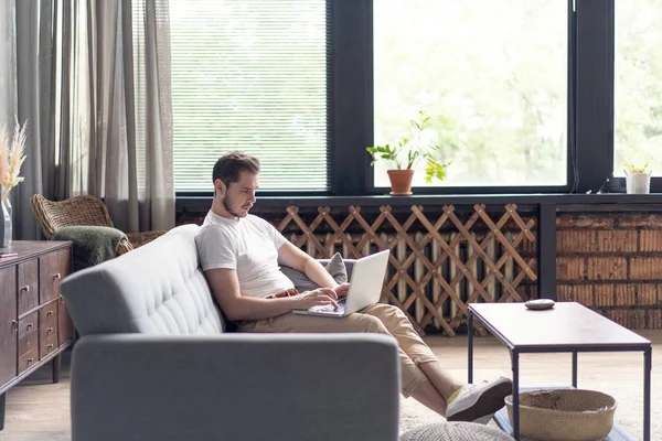 Escribiendo nuevo post de blog. Joven guapo usando su portátil con sonrisa mientras está sentado en el sofá en casa. — Foto de Stock