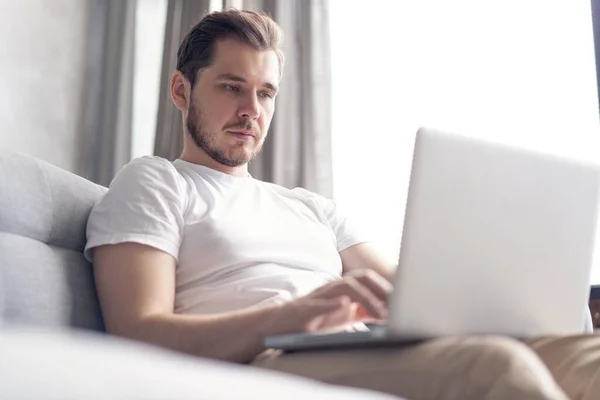 Typing new blog post. Handsome young man using his laptop with smile while sitting on the couch at home.