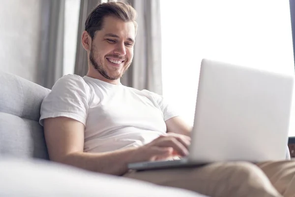 Typing new blog post. Handsome young man using his laptop with smile while sitting on the couch at home.