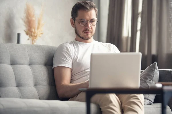 Typing new blog post. Handsome young man using his laptop with smile while sitting on the couch at home.