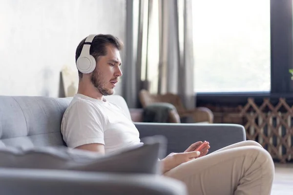 Mi piace la sua musica preferita. Allegro giovane uomo in cuffia che ascolta la musica mentre siede sul suo divano a casa. — Foto Stock