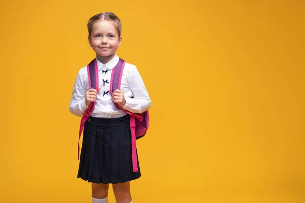 Smiling little cute school girl in uniform with school backpack, isolated on yellow background — Stock Photo, Image