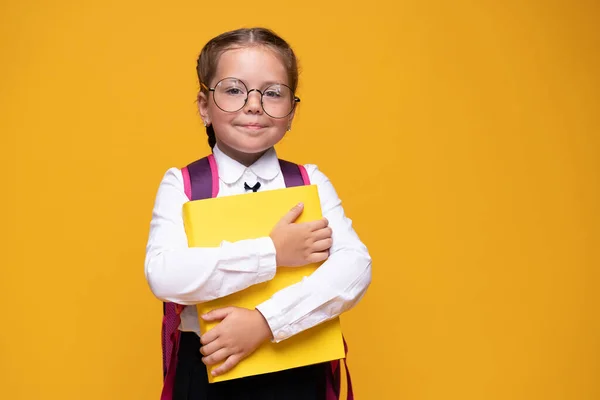 Χαμογελαστό μικρό χαριτωμένο κορίτσι του σχολείου με στολή με σχολική τσάντα, απομονωμένη σε κίτρινο φόντο — Φωτογραφία Αρχείου