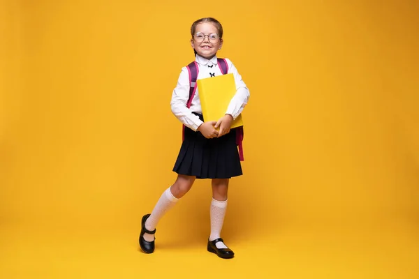 Sorrindo pouco bonito menina da escola em uniforme com mochila escolar, isolado no fundo amarelo — Fotografia de Stock