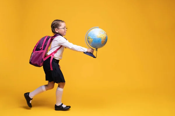 Маленька дівчинка в шкільній формі тримає глобус в руках на жовтому тлі . — стокове фото