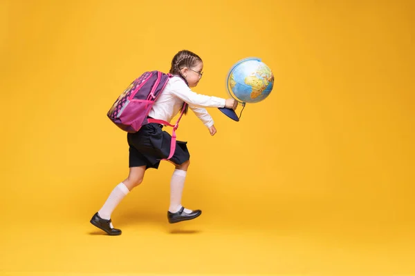 En liten jente i skoleuniform holder en globus i hendene på gul bakgrunn.. – stockfoto