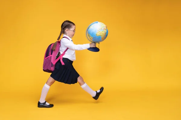 Uma menina em uniforme escolar segura um globo em suas mãos em um fundo amarelo. — Fotografia de Stock
