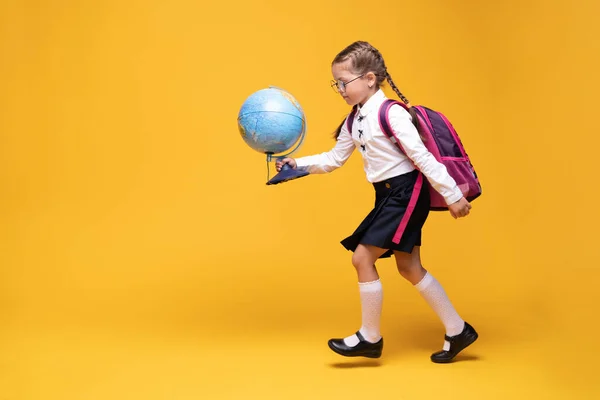 Ein kleines Mädchen in Schuluniform hält auf gelbem Hintergrund eine Weltkugel in den Händen. — Stockfoto