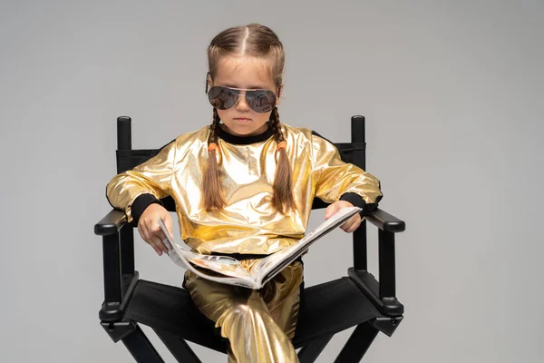 Une petite fille en costume doré s'assoit sur une chaise et lit un livre intéressant. Isolé sur fond jaune. — Photo