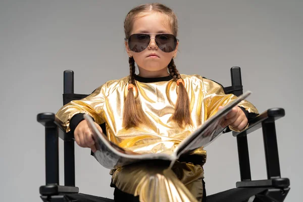 Ein kleines Mädchen im goldenen Anzug sitzt auf einem Stuhl und liest ein interessantes Buch. Vereinzelt auf gelbem Hintergrund. — Stockfoto