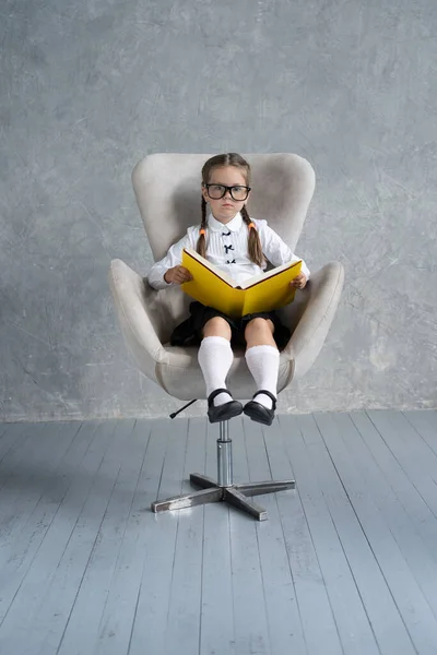 Školačka zaměřená na čtení knihy sedí v křesle. — Stock fotografie