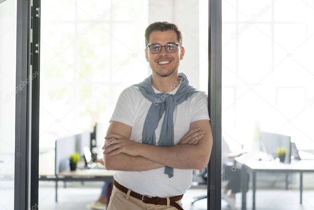 Happy businessman standing in the office with coworkers in the background working by the desk.