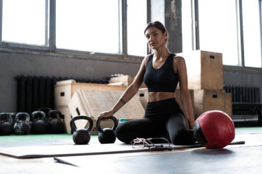 Genç bir kadın antrenmandan sonra yerde oturuyor ve aşağıya bakıyor. Kadın sporcu fitness eğitiminden sonra dinleniyor