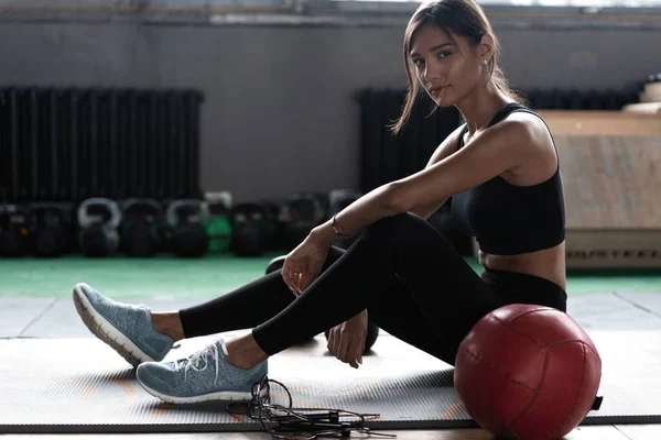 Genç bir kadın antrenmandan sonra yerde oturuyor ve aşağıya bakıyor. Kadın sporcu fitness eğitiminden sonra dinleniyor — Stok fotoğraf