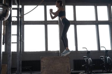 Formda bir spor salonunda atlayan formda genç bir kadın. Kadın sporcu spor salonunda kutu atlayışı yapıyor.