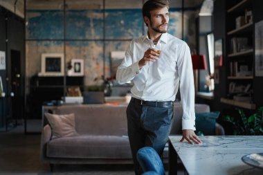 Genç yakışıklı işadamı dikilip kendi ofisinde bir bardak viski tutuyor..