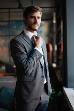 Takım elbiseli şık genç sakallı adam pencerenin yanında dikiliyor ve uzaklara bakıyor.