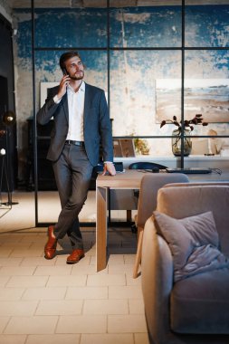 Modern ofiste takım elbiseli yakışıklı bir işadamı ayakta duruyor ve akıllı telefondan konuşuyor..