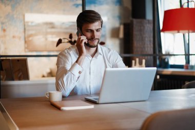 Dizüstü bilgisayarda çalışan ve ofisinde çalışırken cep telefonuyla konuşan kendinden emin genç bir adam.