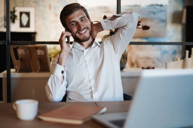 Dizüstü bilgisayarda çalışan ve ofisinde çalışırken cep telefonuyla konuşan kendinden emin genç bir adam.