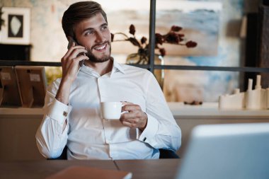 Dizüstü bilgisayarda çalışan ve ofisinde çalışırken cep telefonuyla konuşan kendinden emin genç bir adam.