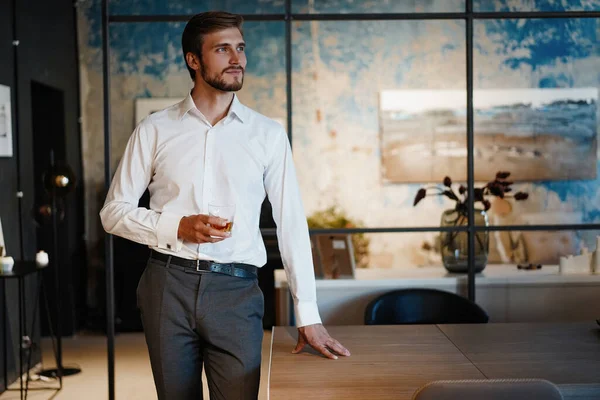 Jovem empresário bonito se levantar e segurar um copo de uísque em seu próprio escritório. — Fotografia de Stock