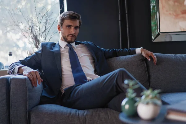 若く魅力的なビジネスマンは彼の近代的なオフィスでソファに座っている間、ビジネスについて考えています. — ストック写真