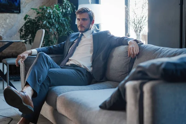 若く魅力的なビジネスマンは彼の近代的なオフィスでソファに座っている間、ビジネスについて考えています. — ストック写真
