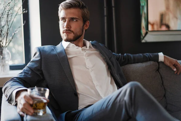 Bonito e bem sucedido empresário em terno elegante segurando uísque de vidro enquanto sentado no escritório. — Fotografia de Stock