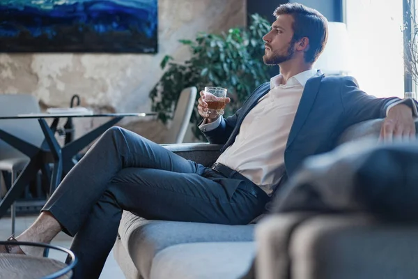 Przystojny i odnoszący sukcesy biznesmen w stylowym garniturze trzymający szklaną whisky podczas siedzenia w biurze. — Zdjęcie stockowe