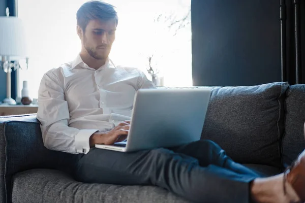 Multitasking. Bello giovane uomo che lavora con il computer portatile mentre è seduto sul divano in ufficio — Foto Stock