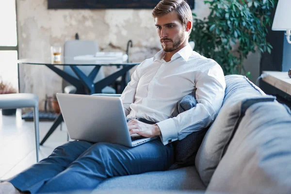 Multitasking. Schöner junger Mann arbeitet mit Laptop, während er auf der Couch im Büro sitzt — Stockfoto