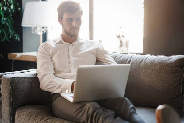 Multitasking. Bello giovane uomo che lavora con il computer portatile mentre è seduto sul divano in ufficio — Foto Stock