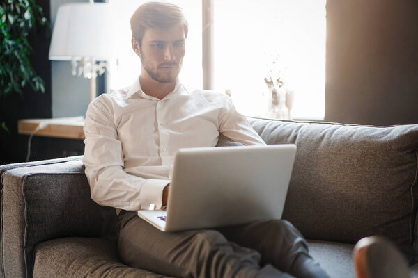 Multitasking. Handsome young man working with laptop while sitting on the couch in office