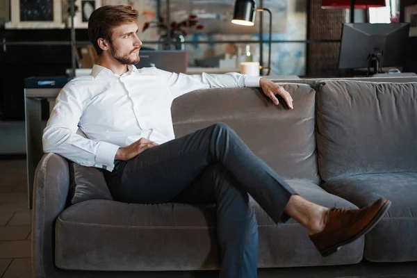 Jonge en charmante zakenman denkt na over zaken terwijl hij op de bank zit in zijn moderne kantoor. — Stockfoto