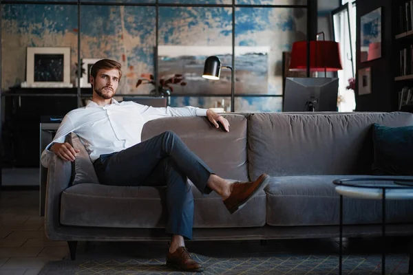 Молодой и очаровательный бизнесмен думает о бизнесе, сидя на диване в своем современном офисе. — стоковое фото