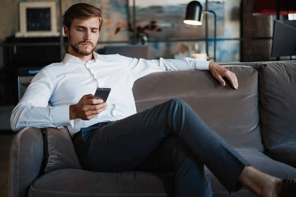 笑顔ひげを生やした男作りテキストメッセージ上のスマートフォンでソファに座っている間現代のオフィス. — ストック写真