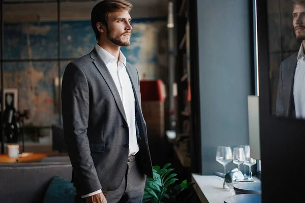 Homem barbudo jovem elegante em um terno está parado perto da janela e olhando para longe — Fotografia de Stock