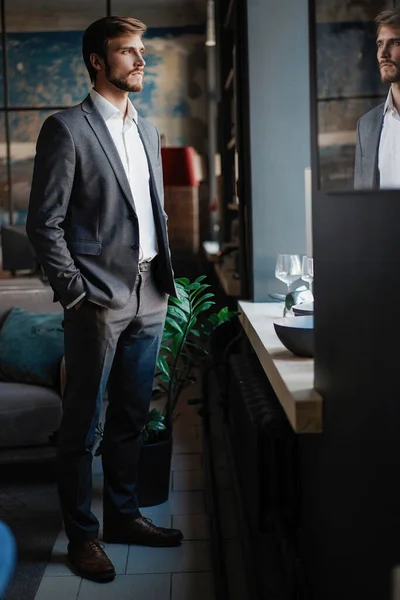 Stylish young bearded man in a suit is standing near the window and looking far — Stock Photo, Image