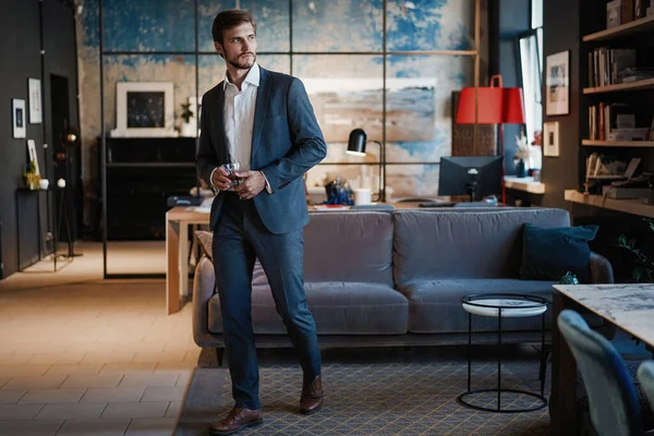 Młody przystojny biznesmen stoi i trzyma szklankę whisky we własnym biurze. — Zdjęcie stockowe