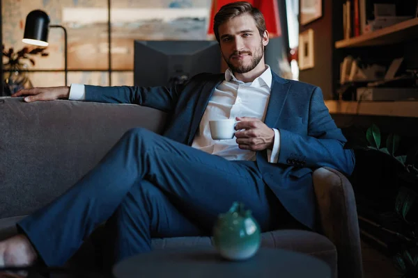Bonito jovem segurando xícara de café sentado no sofá no escritório — Fotografia de Stock